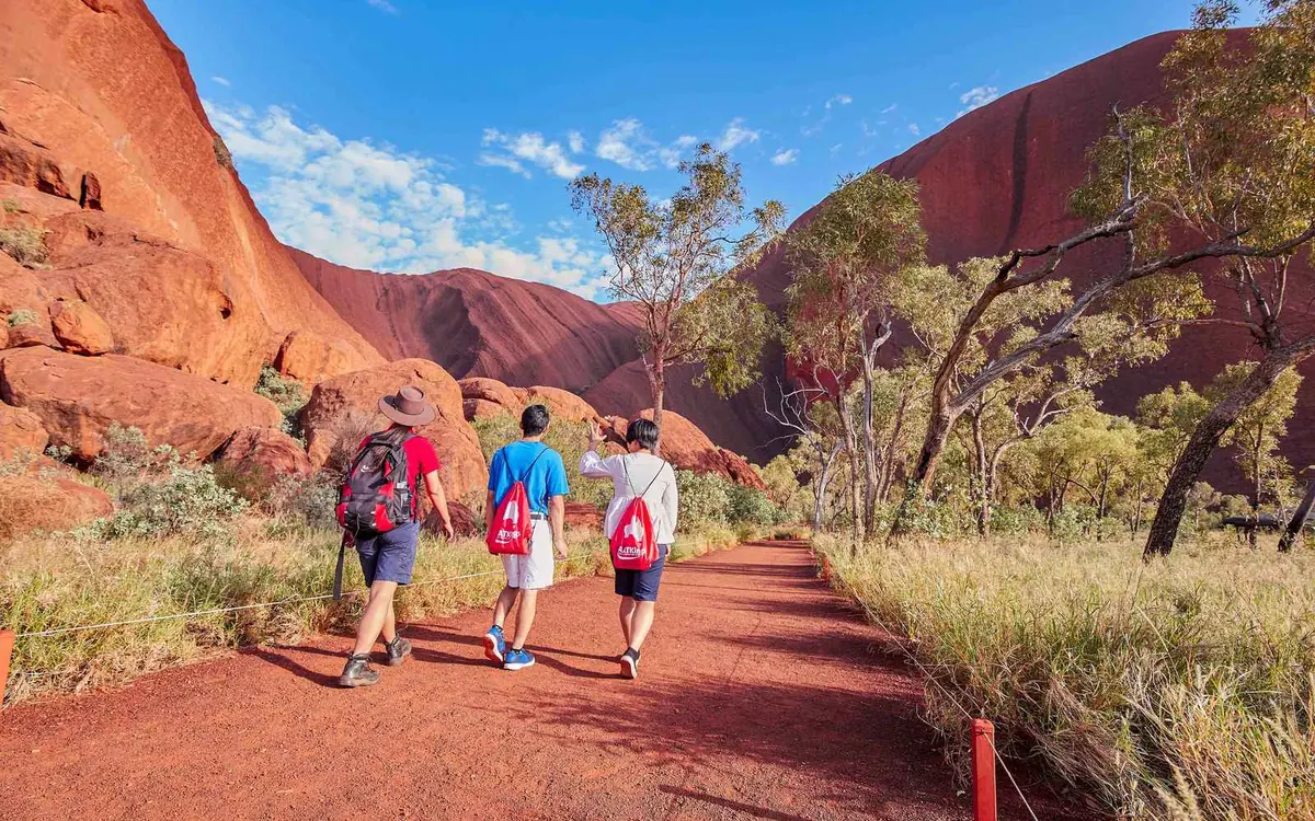 A guided group on the red sand at the foot of Uluru, spinifex and desert oaks in the foreground and the rock rising behind them