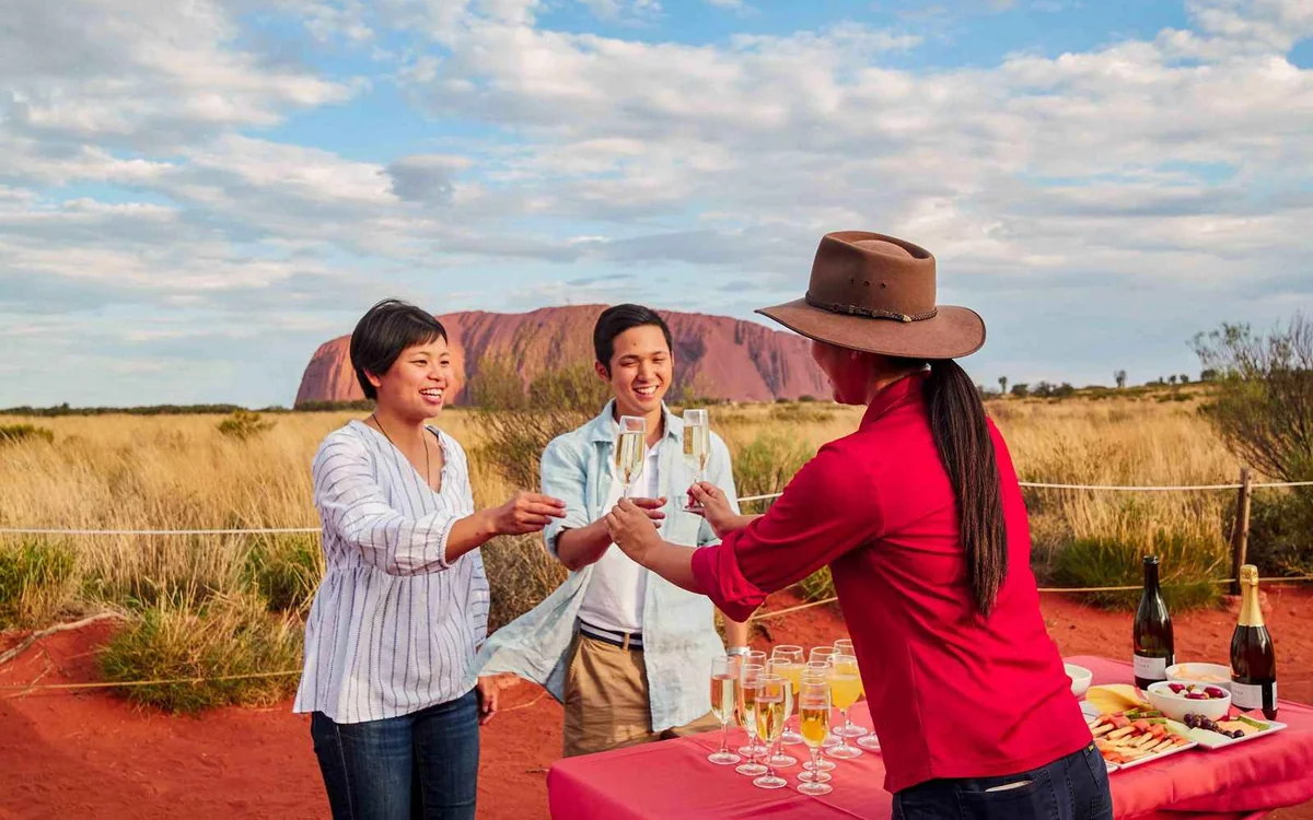 A small guided group standing with their guide on the red sand below the flank of Uluru, listening rather than photographing