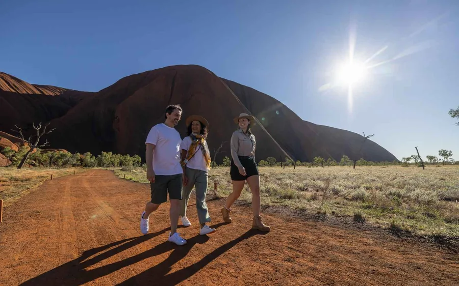Walkers on a sandy trail following the foot of the red rock face of Uluru