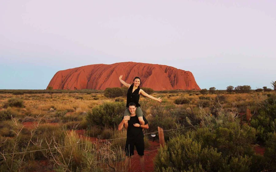 Uluru seen across the red desert plain from the road, low scrub in the foreground