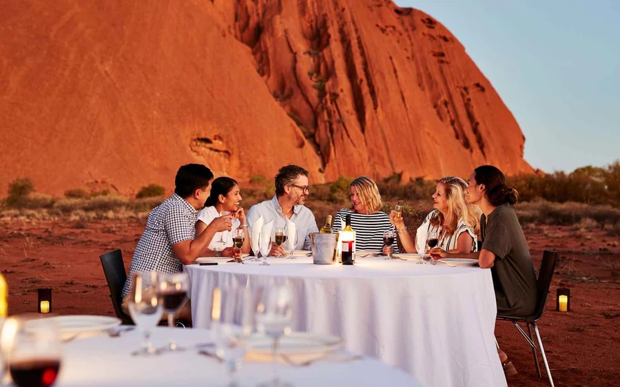 A table laid for dinner in the desert at dusk with the outline of Uluru behind