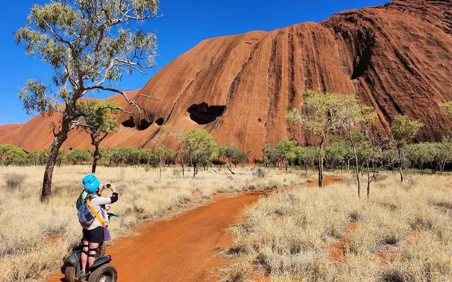 A person on a Segway on a sandy track with the red rock of Uluru behind